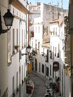 Carrer de la Carreta, Sitges: a narrow whitewashed old-town lane with iron wall lanterns and potted plants sloping downhill