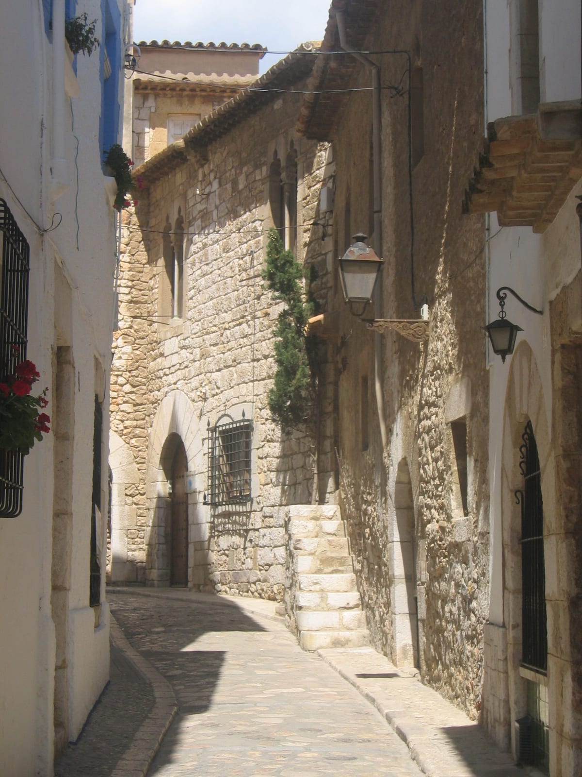 Carrer d’en Bosc, Sitges: a narrow cobbled old-town lane of honey-coloured stone houses with a gothic doorway and iron lanterns
