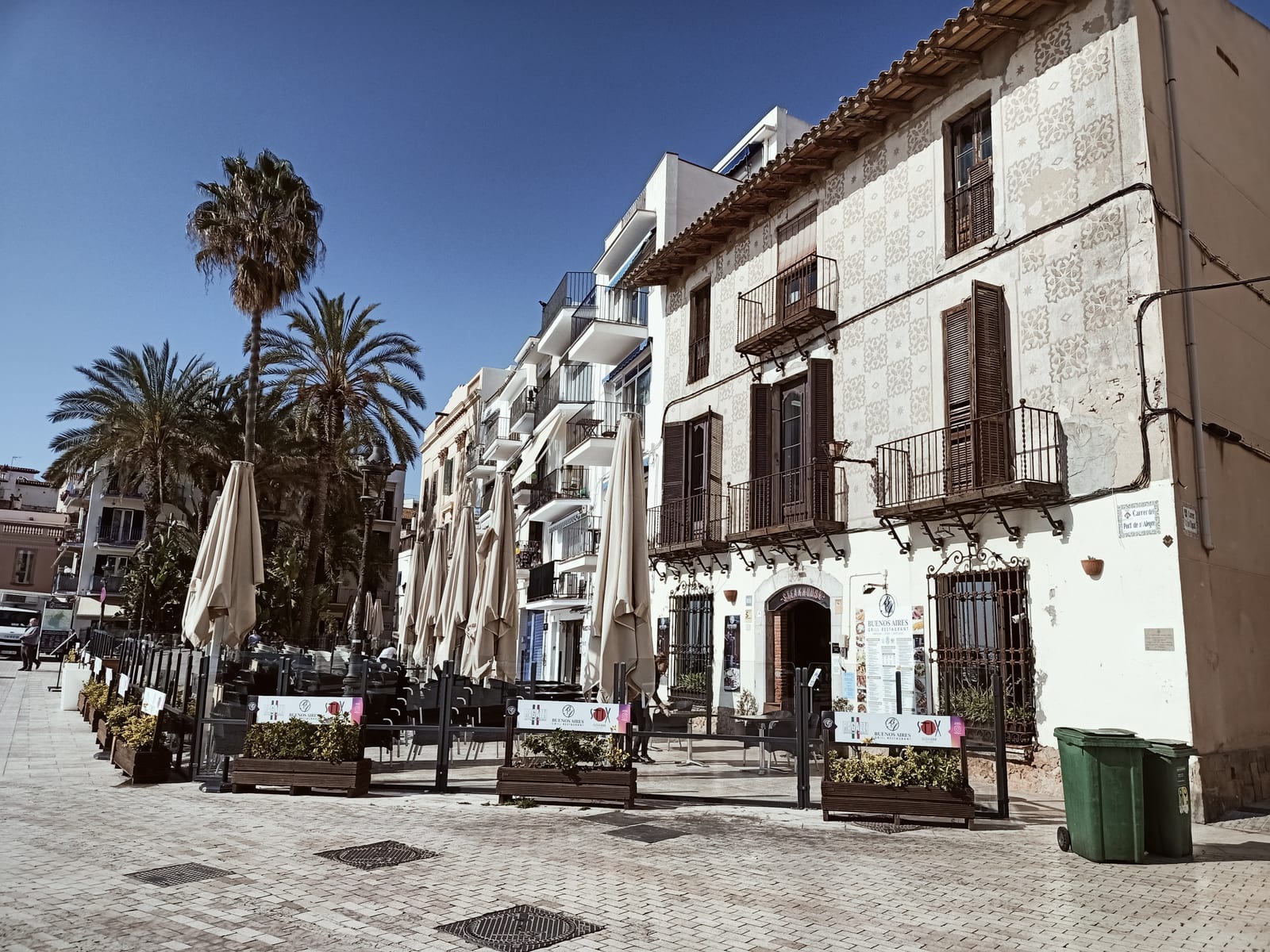 Carrer del Port Alegre, Sitges: a palm-shaded paved square in the old fishermen’s quarter with a ceramic-tiled townhouse façade and café terraces